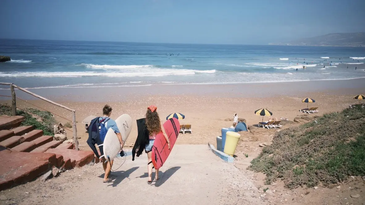 Two girls carrying surfboards down to the beach, with surfers in the water