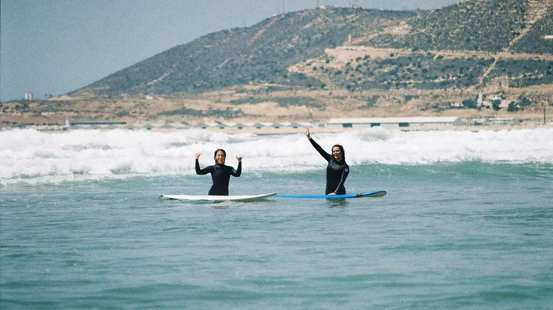 Two girls with their boards in the water waving and smiling, in Morocco